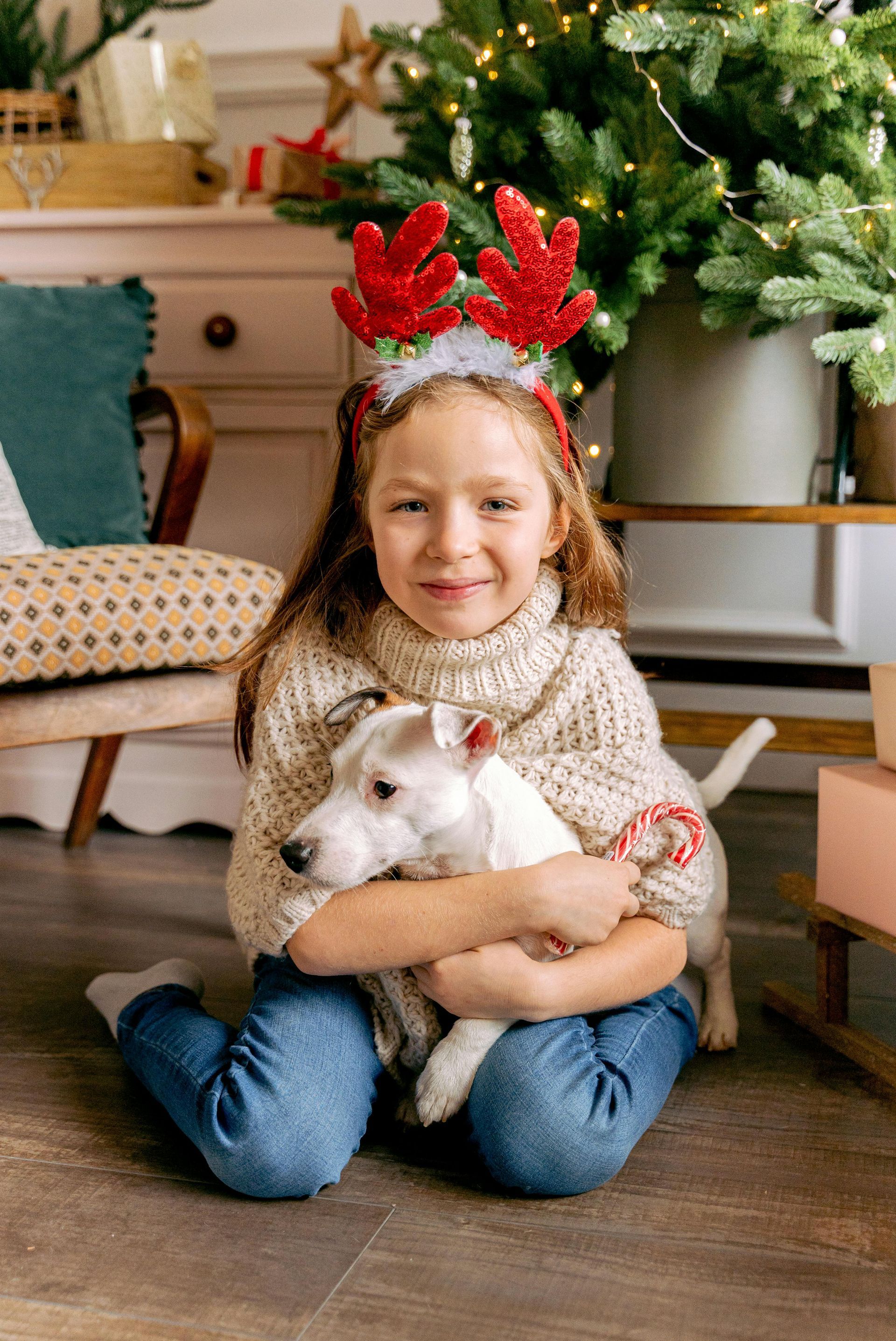 Girl with reindeer antlers hugs a white dog, sitting in front of a Christmas tree.