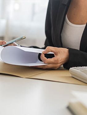 Woman in black blazer examines paperwork at desk with calculator and envelope.