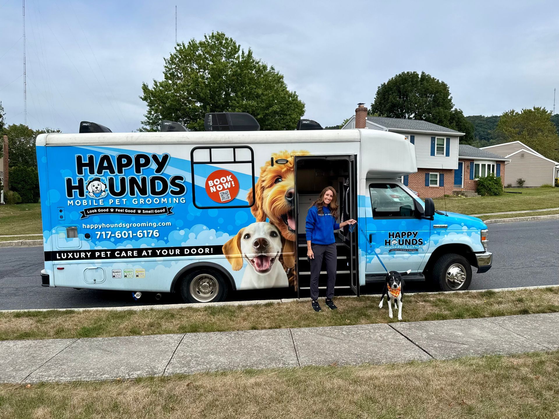 Happy Hounds mobile pet grooming van parked outside with groomer and dog beside the vehicle.