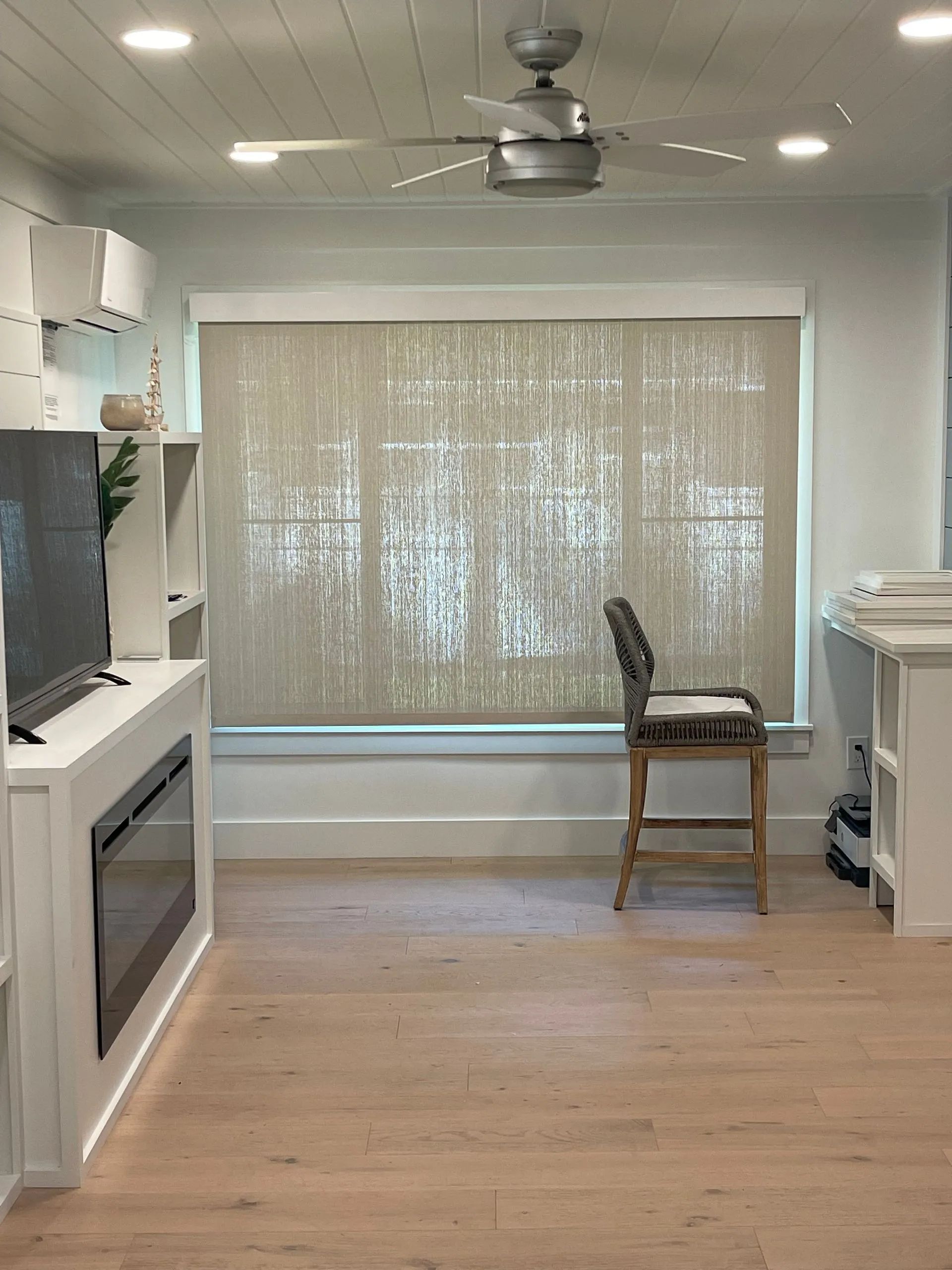 Bright kitchen with a large window, ceiling fan, white cabinets, wood floor, and a stool by the window.