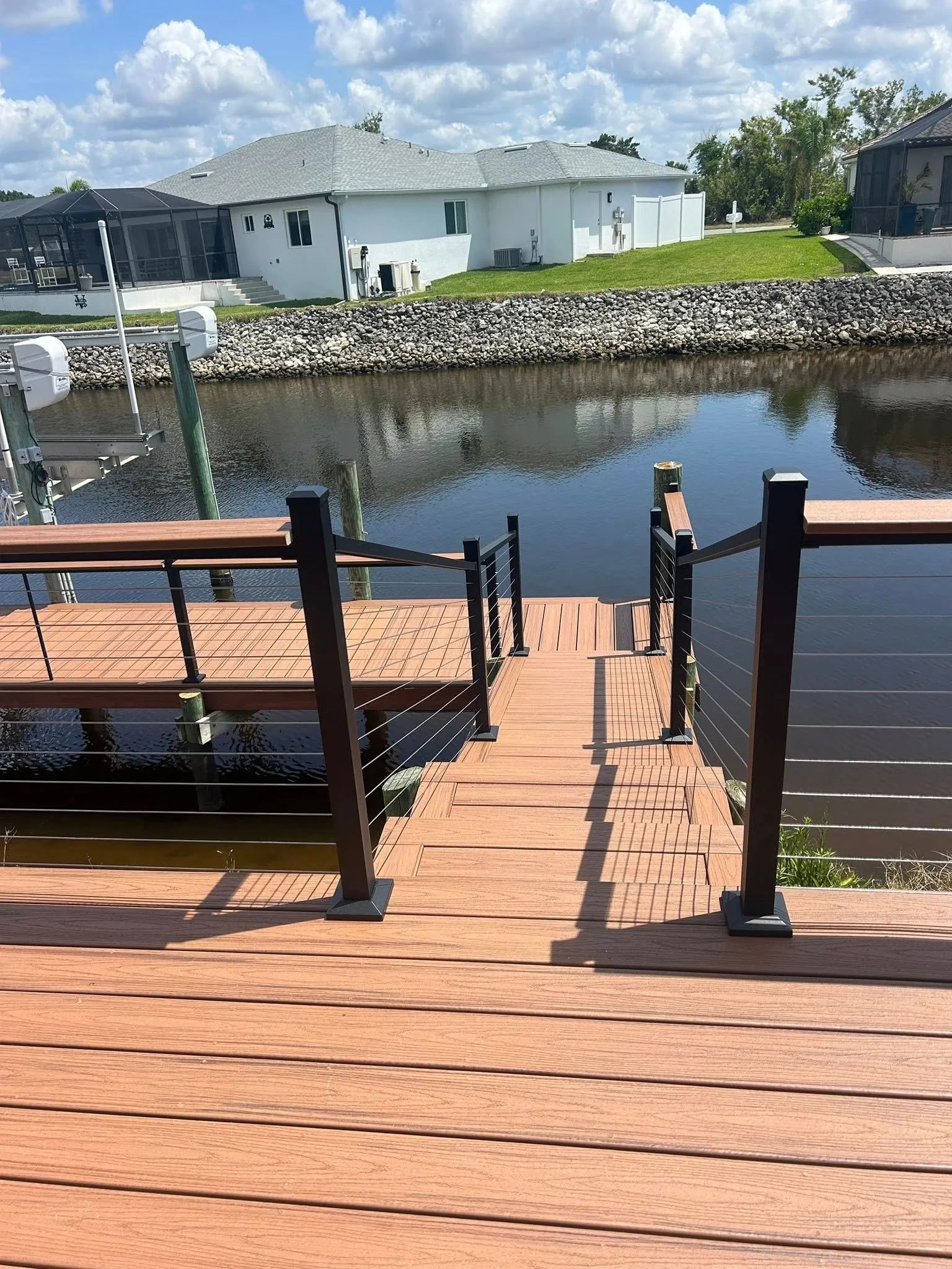 Wooden dock with stairs leading to water, black railings, sunny day, houses in background.