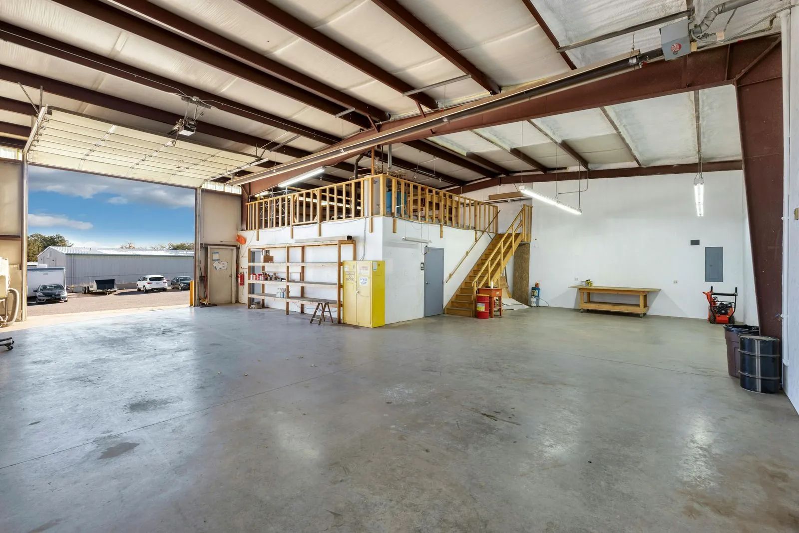 Interior view of a large warehouse with a mezzanine and open bay door. Concrete floor, metal beams, white walls.
