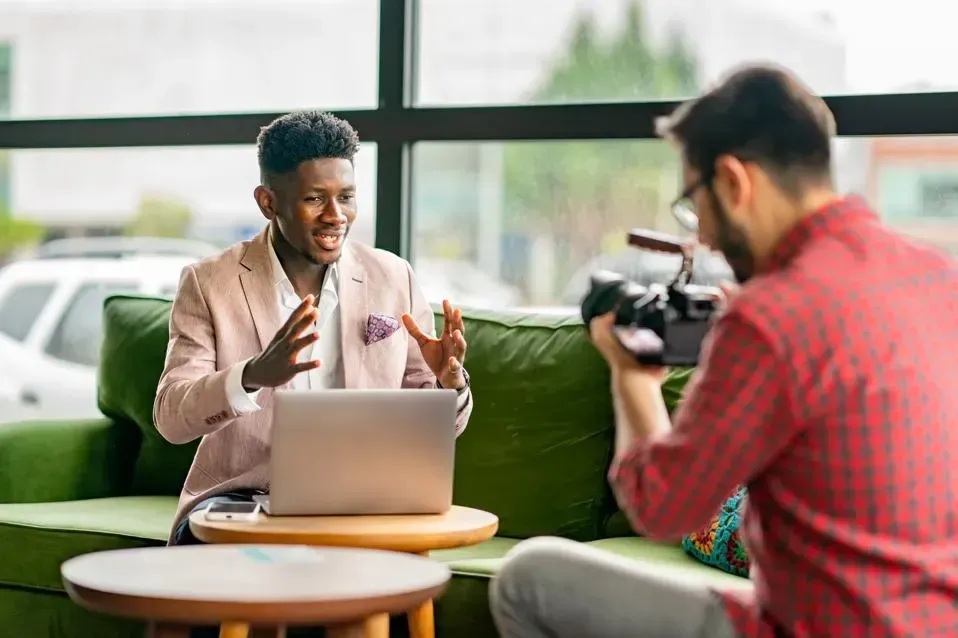 A man is sitting on a couch with a laptop and a camera.