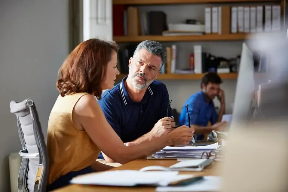 A man and a woman are sitting at a desk in an office talking to each other.