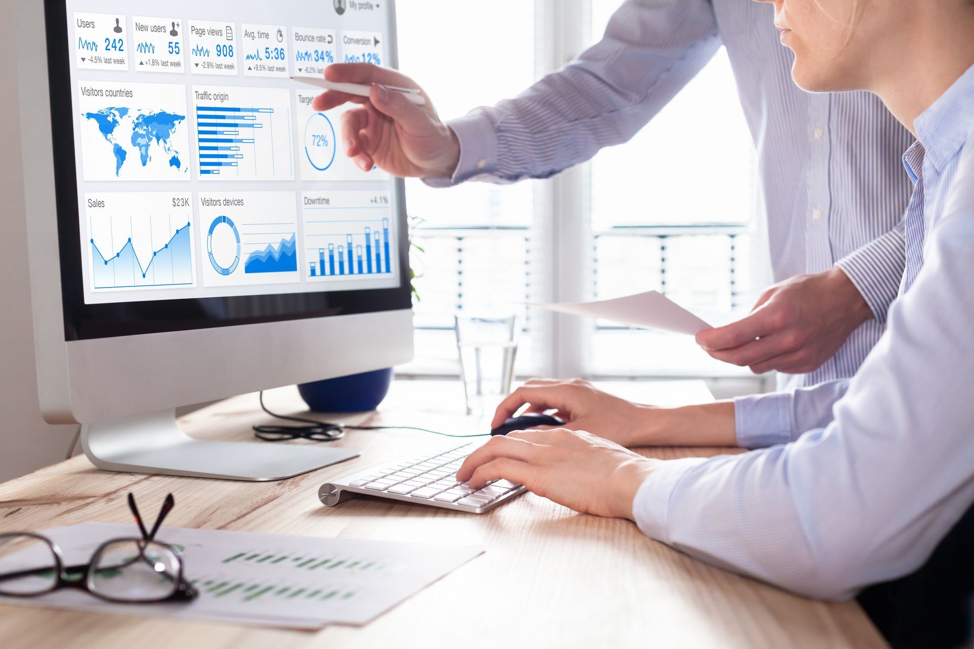 Two coworkers reviewing blue analytics charts on a desktop monitor in a bright office