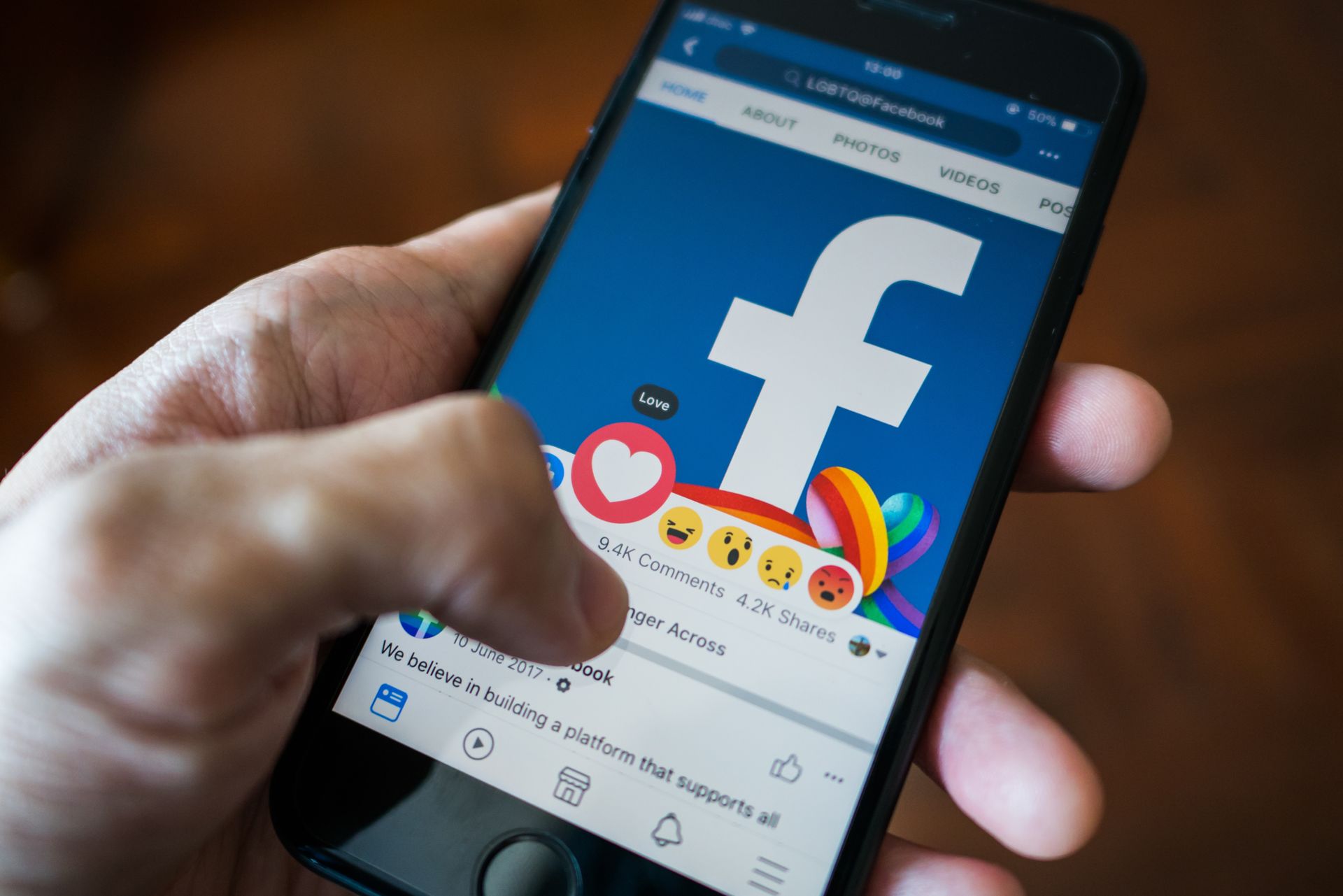 Hand holding a smartphone displaying a Facebook feed with emoji reactions. The screen shows the Facebook logo.