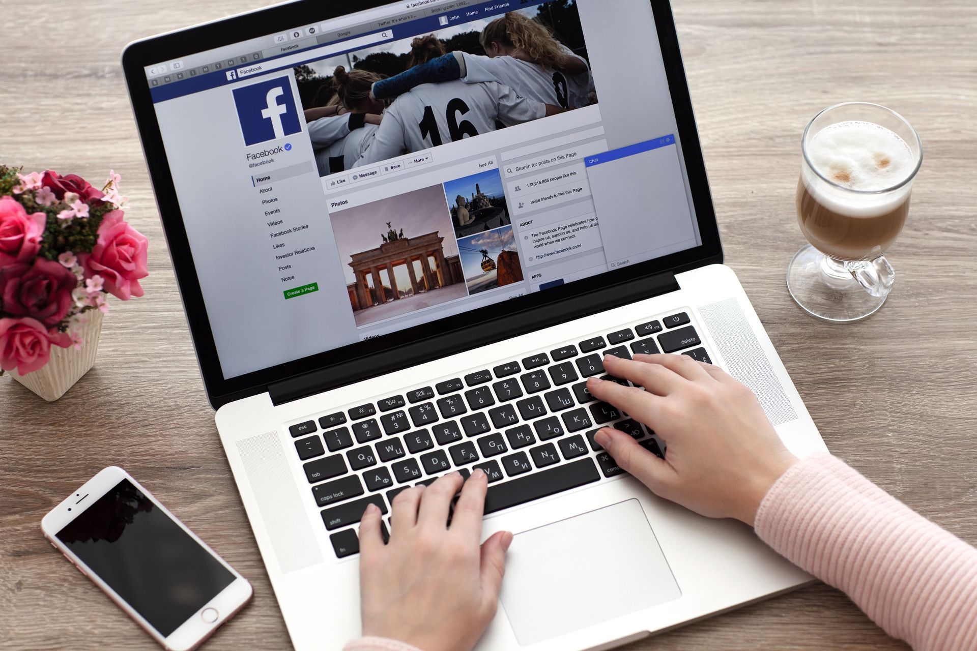 Person using a laptop with Facebook open, typing on the keyboard. A phone, flowers, and a drink sit nearby.