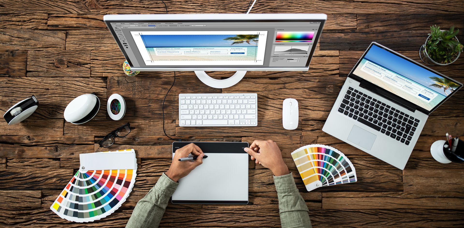 Designer using graphics tablet with a monitor and laptop on a wooden desk with color swatches. Designer using graphics tablet with a monitor and laptop on a wooden desk with color swatches.