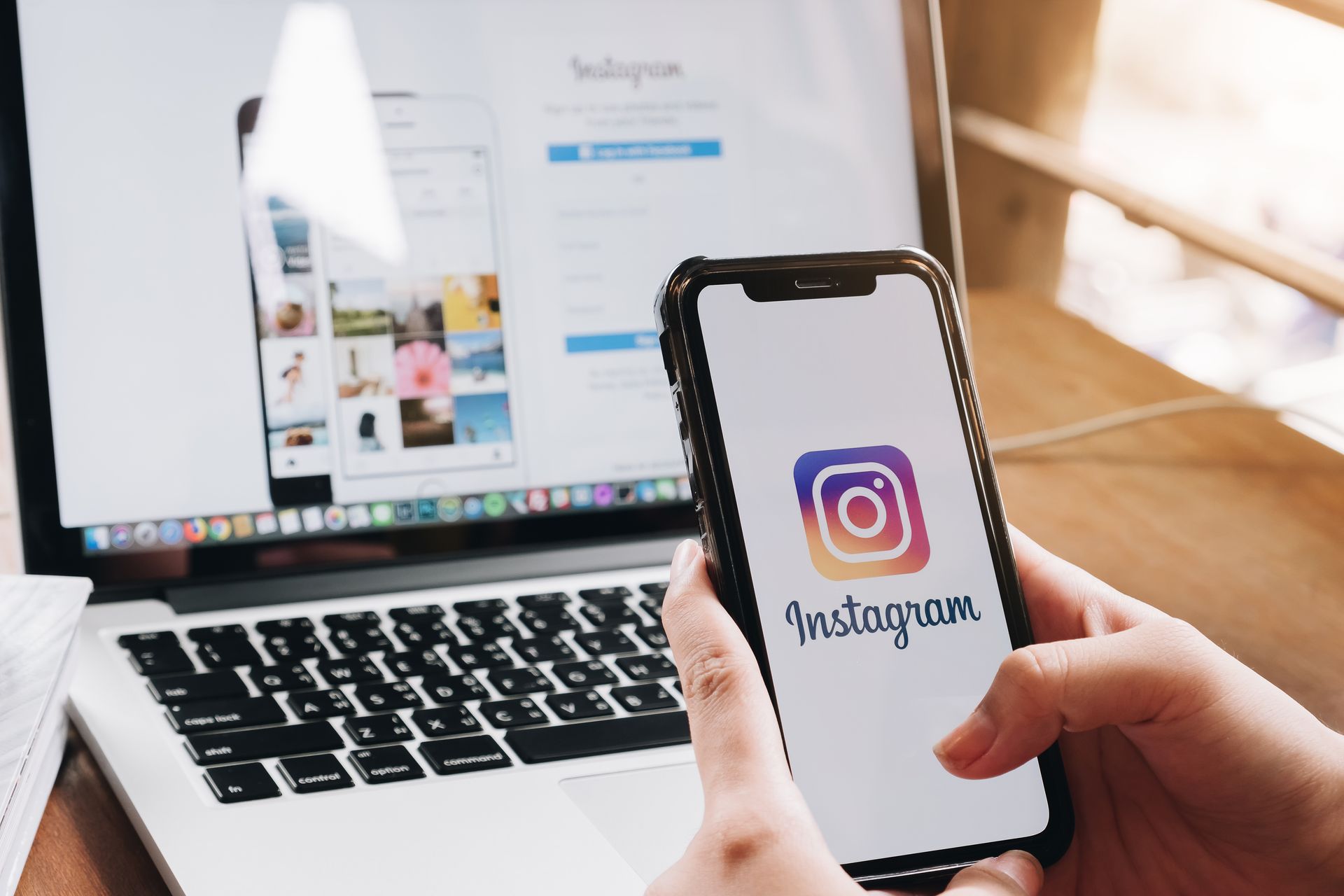 Hands holding a smartphone with Instagram logo in front of a laptop showing the Instagram website