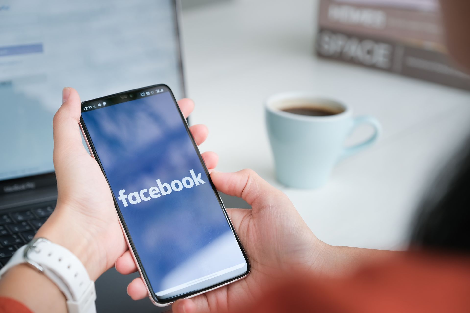 Hands holding a smartphone displaying the Facebook logo. A laptop, mug, and book are in the background.