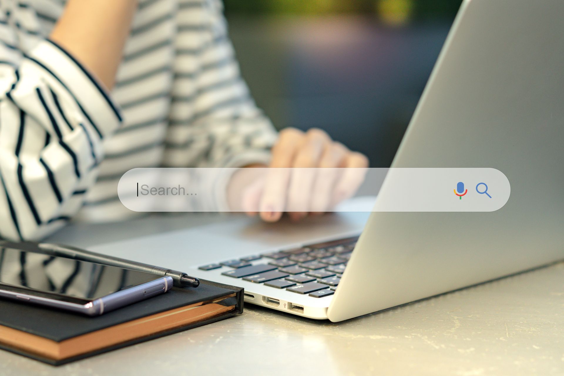 Person typing on a laptop beside a smartphone and notebook, with a search bar overlay on screen