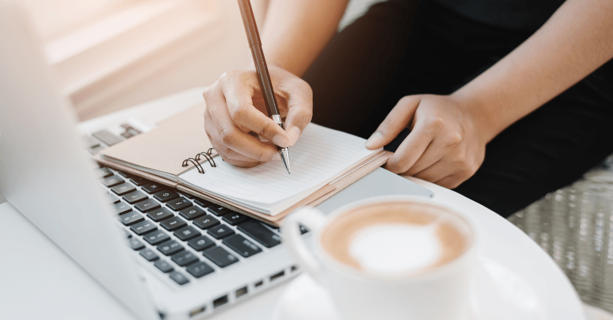 A person is writing in a notebook in front of a laptop computer.