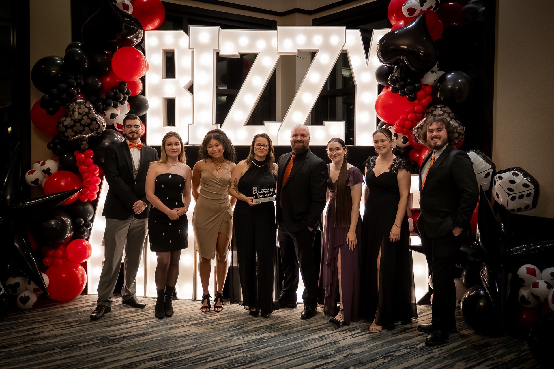 Group of people posing on a red carpet before a lit “RIZZA” backdrop with black and red balloons