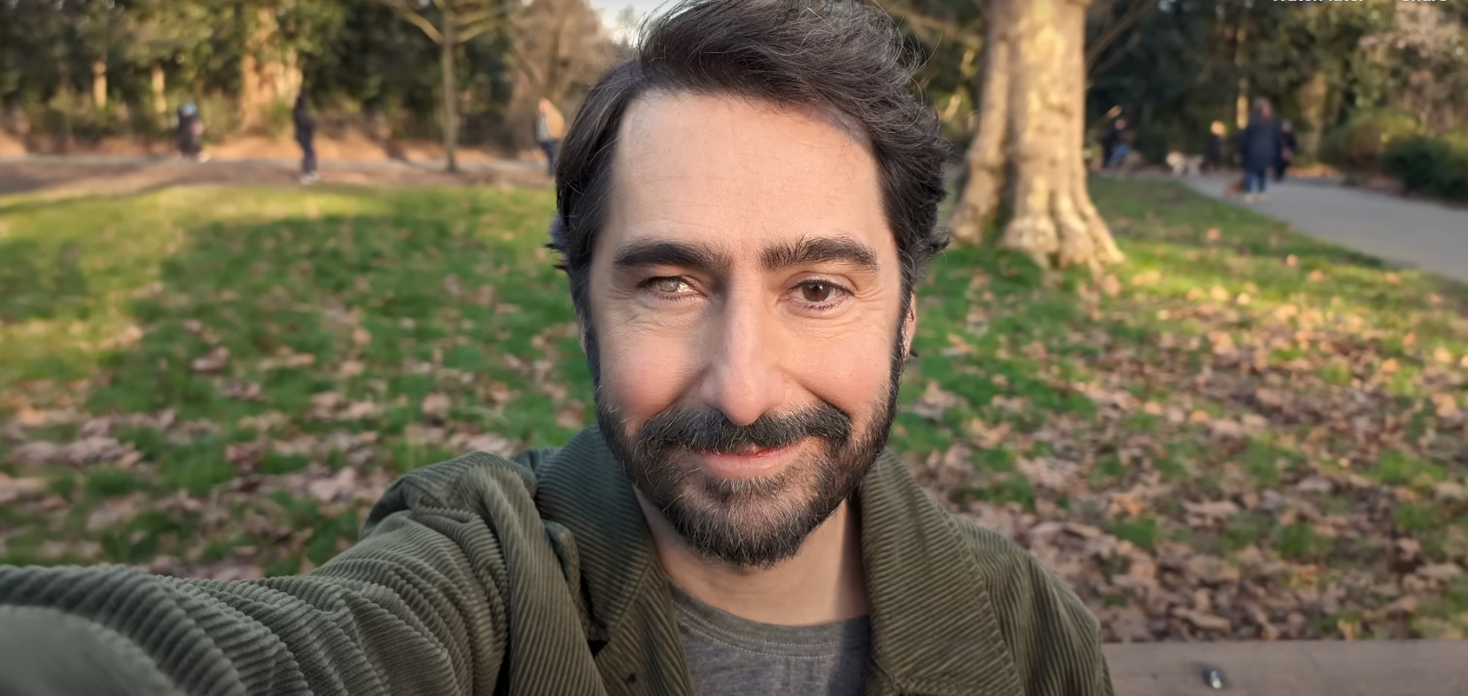 A man with a beard is taking a selfie in a park.