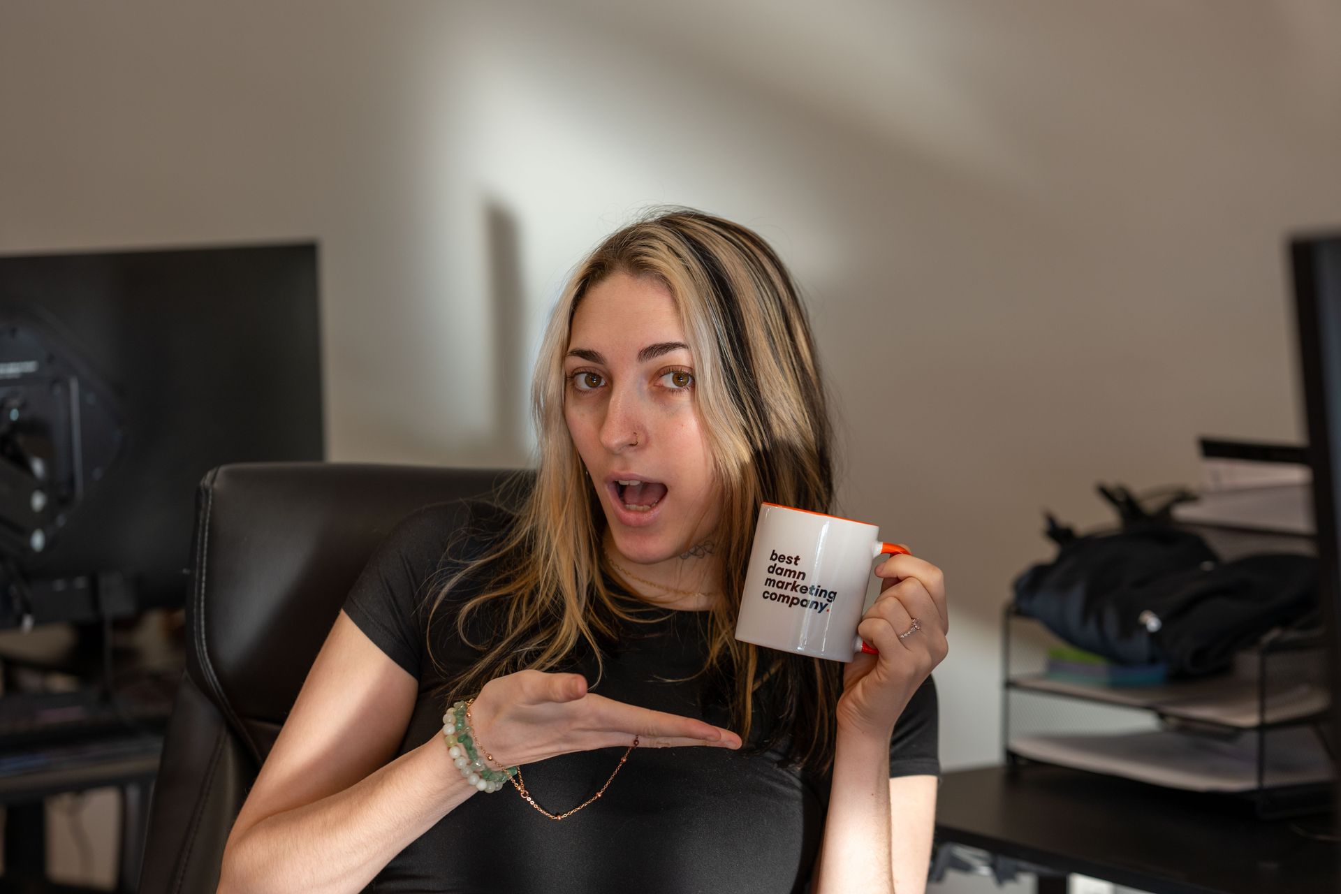 Woman holding a mug and pointing at it, seated at a desk in an office with a surprised expression.