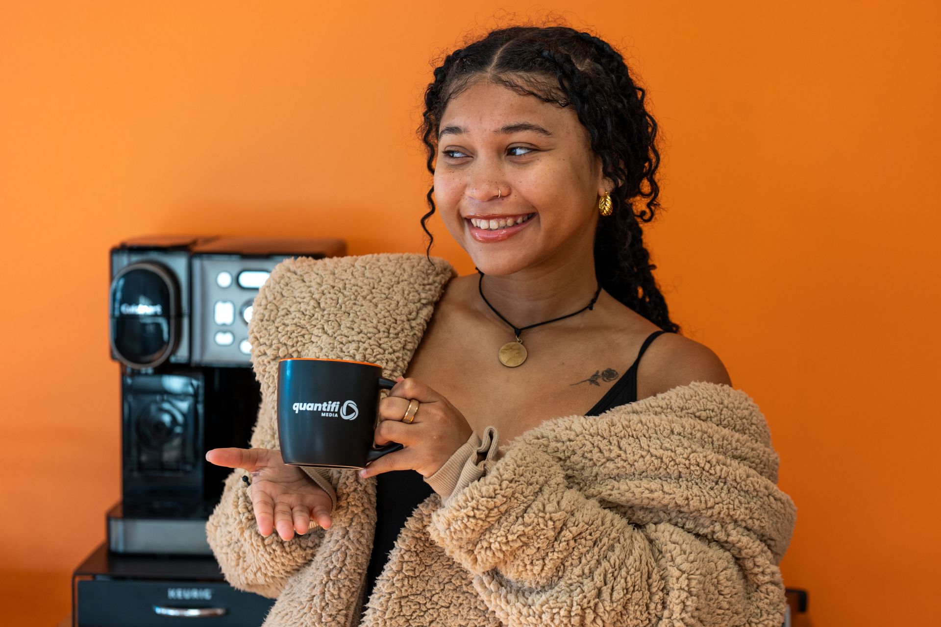 Smiling person holding a mug beside a coffee machine against an orange wall