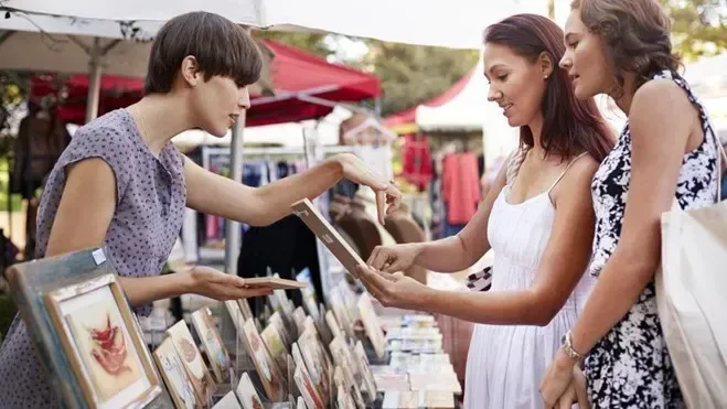 Three women are standing around a table at a market looking at paintings.