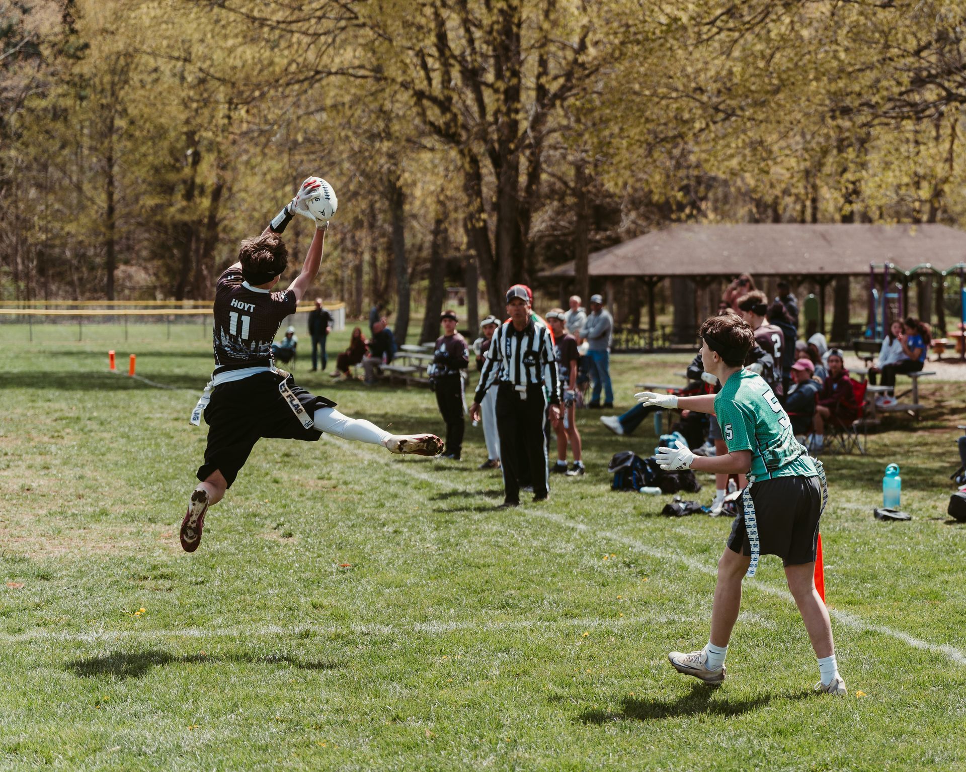 Youth flag football game on a grassy field, player leaping to catch the ball while others watch near the sideline.