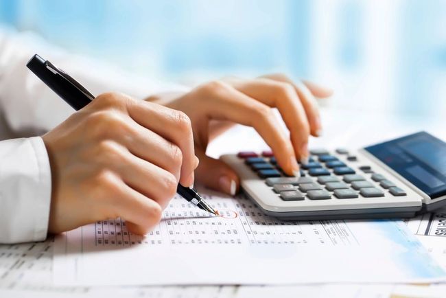 Hands using a calculator and pen on paperwork beside a laptop
