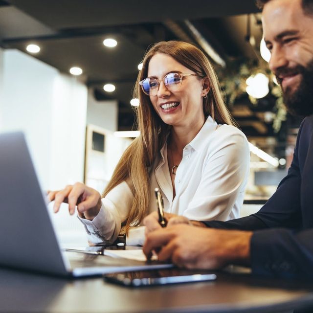 Two coworkers smiling and pointing at a laptop in a modern office setting