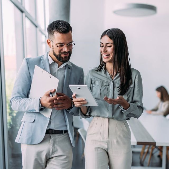 Two coworkers in light business attire reviewing documents on tablets in a bright office hallway