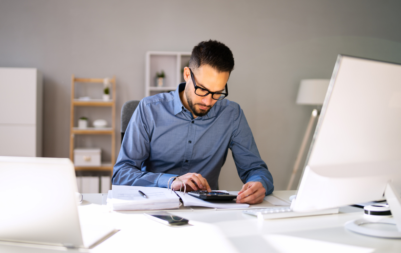 Man in blue shirt working at a desk with laptop and papers in a bright office
