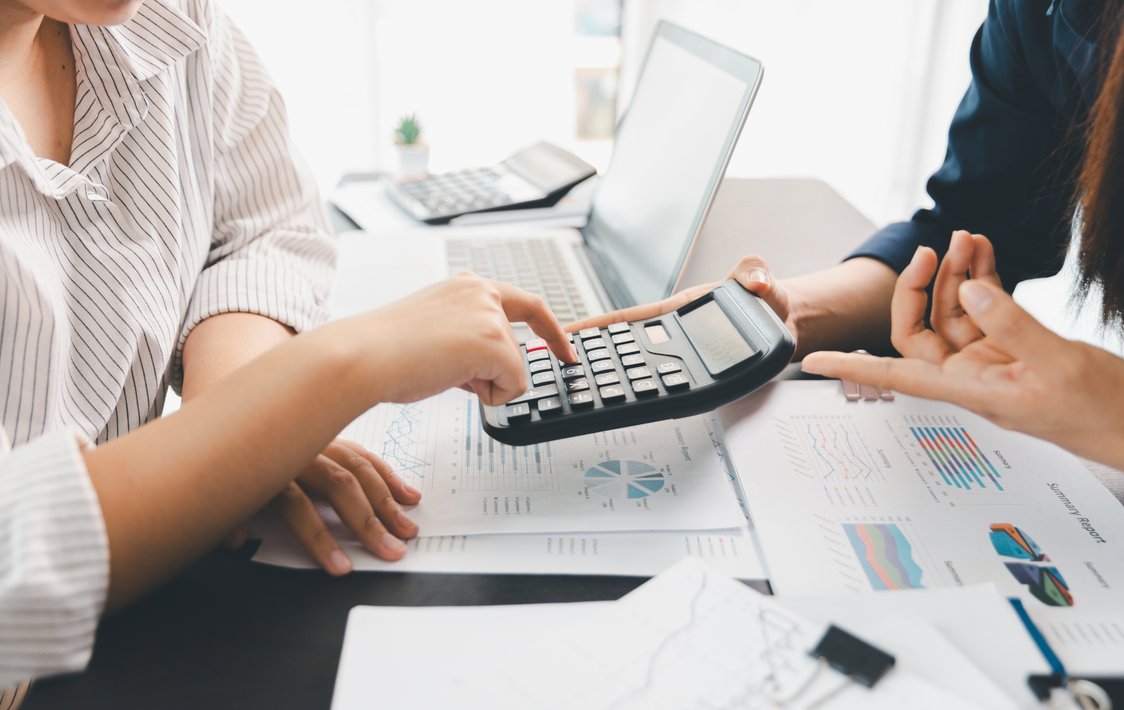 Two people reviewing charts and using a calculator at a desk with a laptop