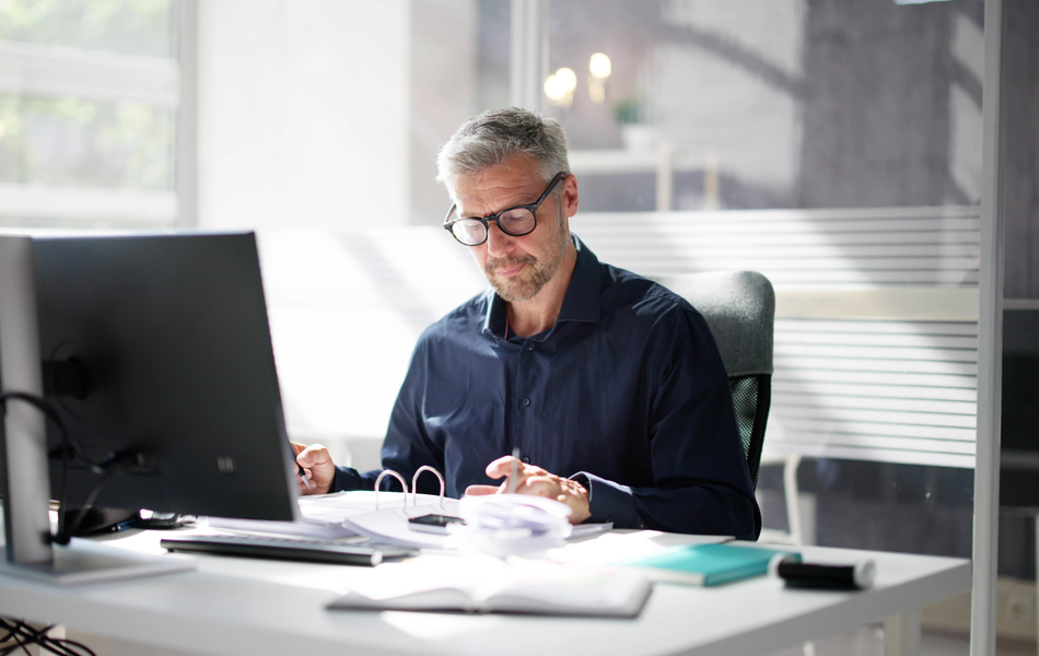 Man writing at a desk in a bright office, with a computer monitor and papers nearby.