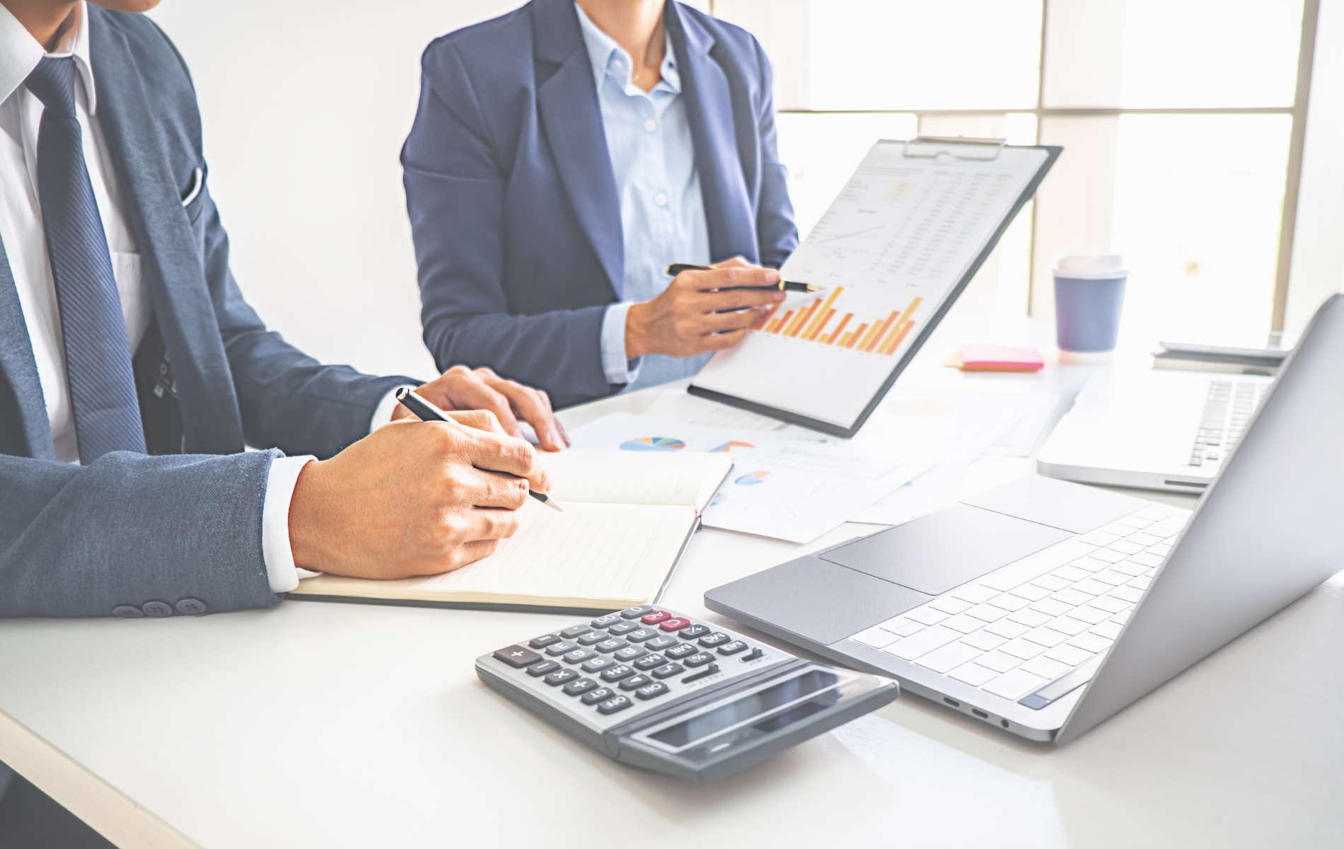 Two businesspeople reviewing charts at a bright office desk with a laptop and calculator.