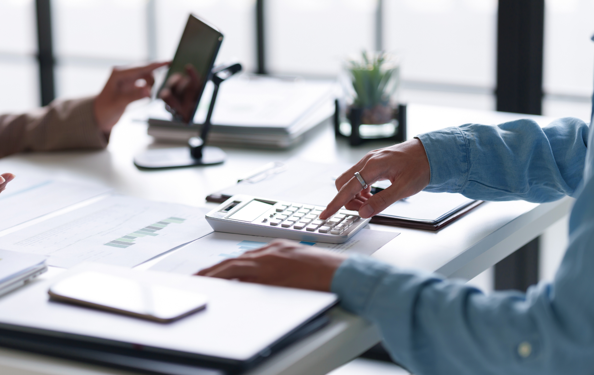 Hands typing on a laptop at a bright office desk with a phone on a stand and papers nearby