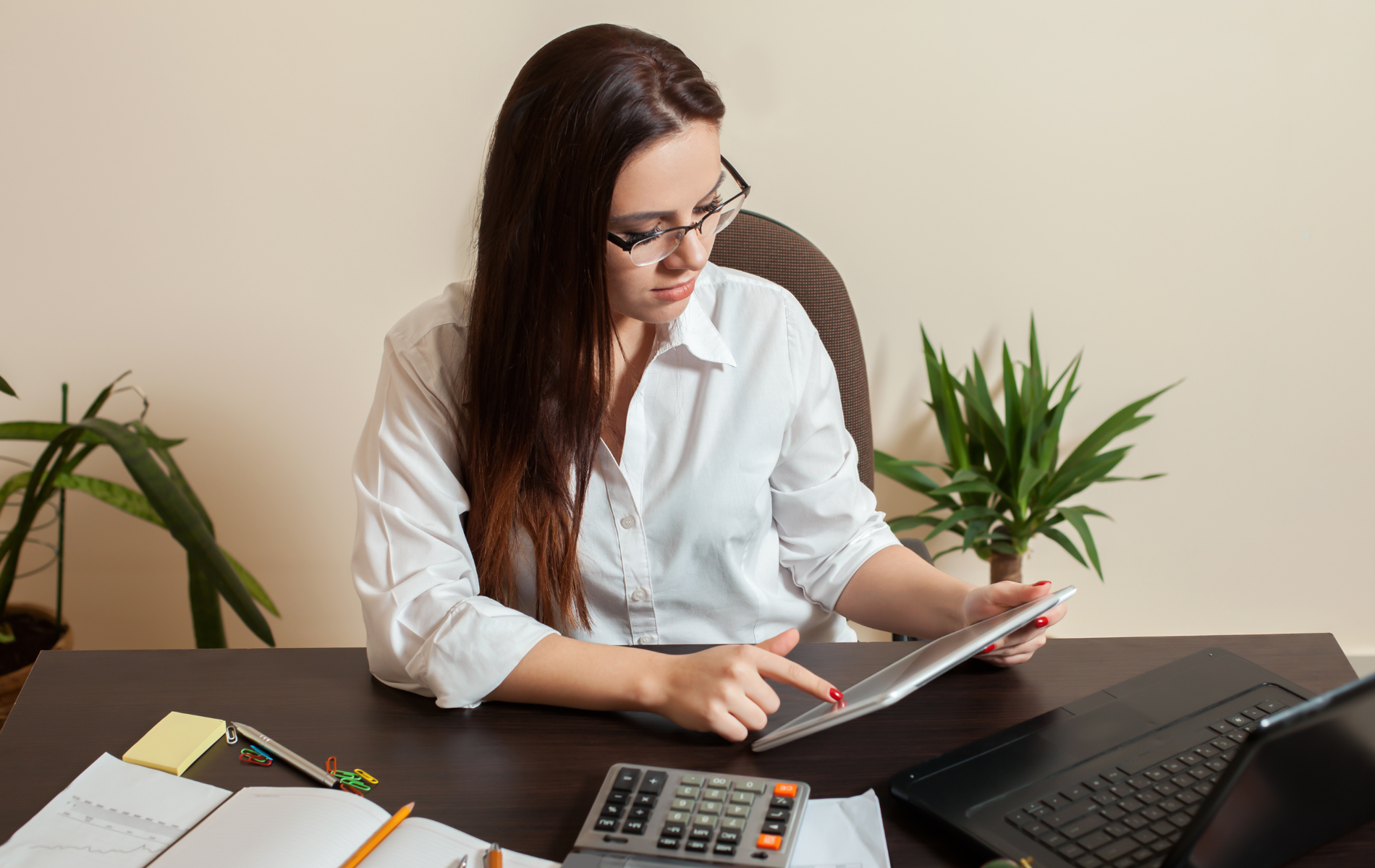 Woman in white shirt using a tablet at a desk with calculator, laptop, and papers in an office