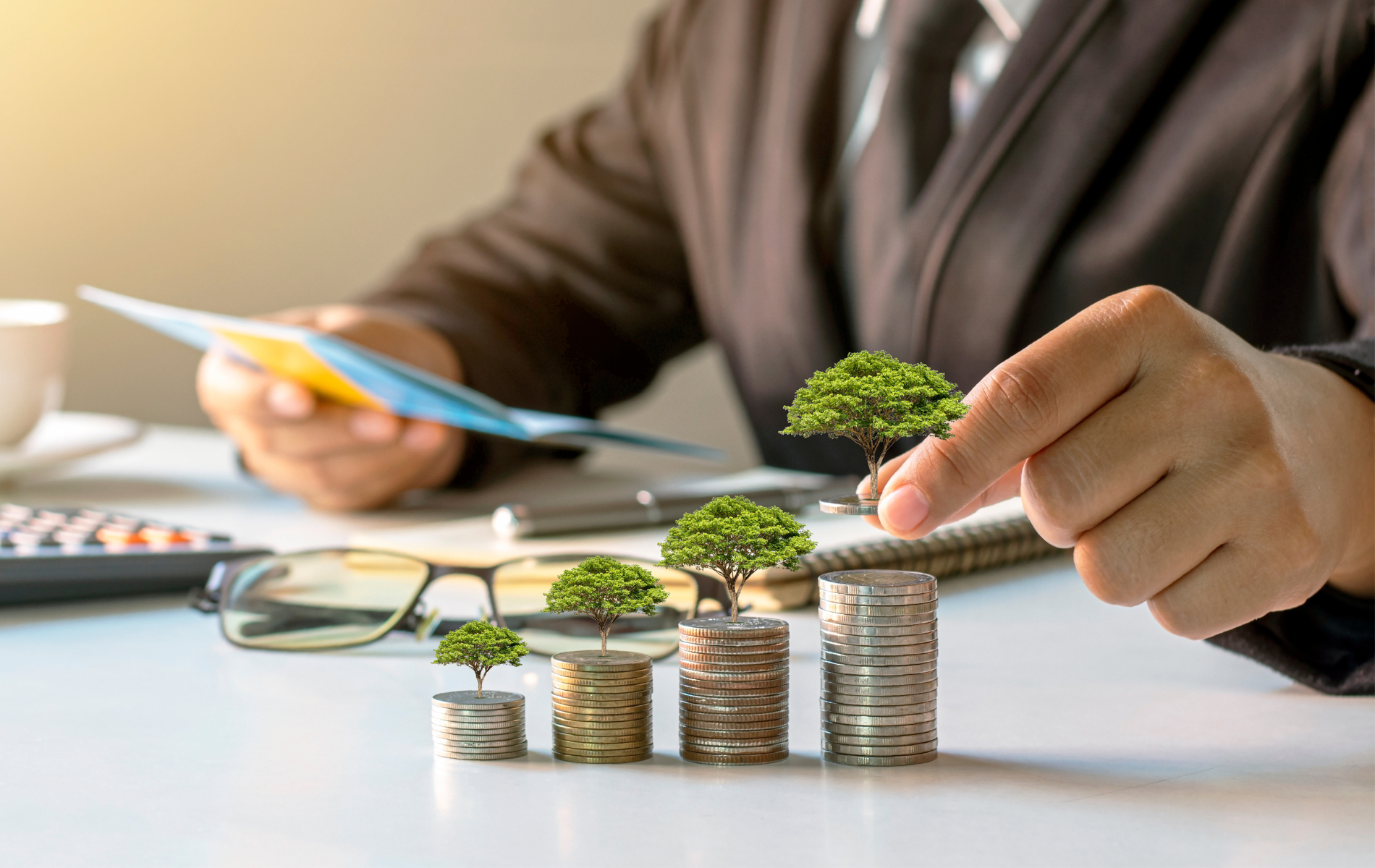 Hands arranging coin stacks with tiny trees, calculator and glasses on desk, symbolizing green investment