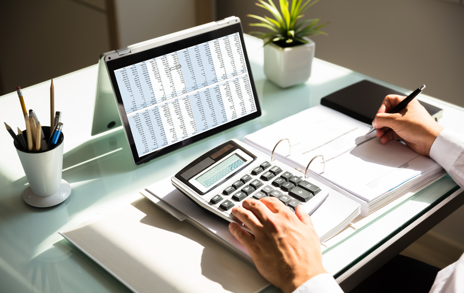 Hands using a calculator beside a spreadsheet notebook and pen on a tidy desk with a tablet and plant