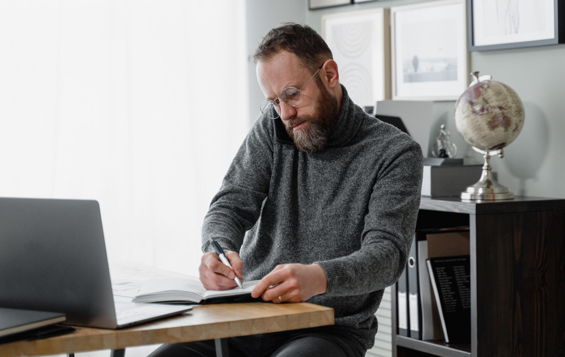 Man writing at a desk beside a laptop in a bright office, with a globe on a shelf behind him