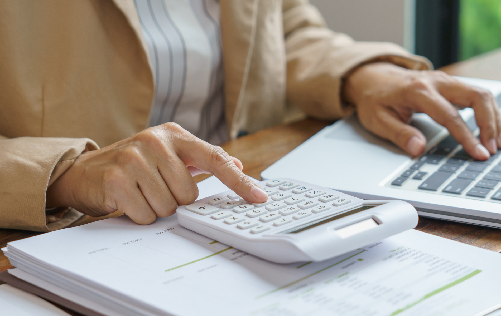 Person using a calculator beside a laptop on a desk with paperwork