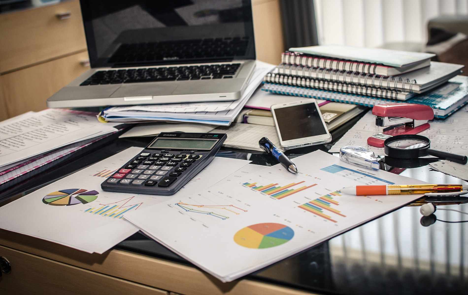 Office desk with laptop, calculator, notebooks, phone, and charts spread across papers and pens