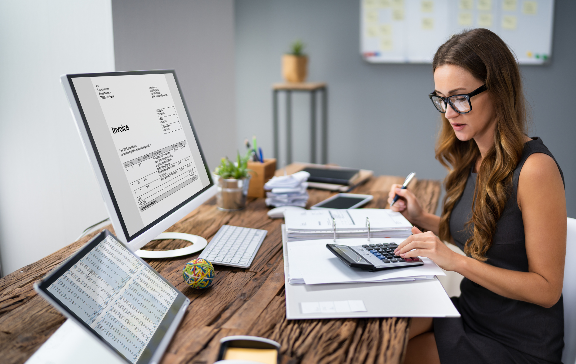 Woman working at a desk with laptop, papers, calculator, and desktop monitor in a bright office