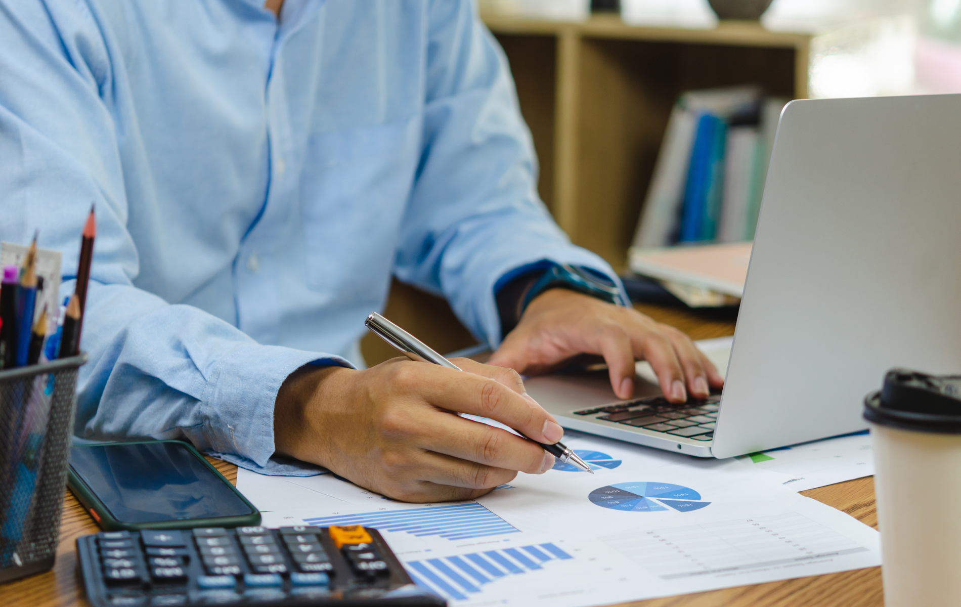 Person in a blue shirt using a laptop and calculator at a desk with financial charts.