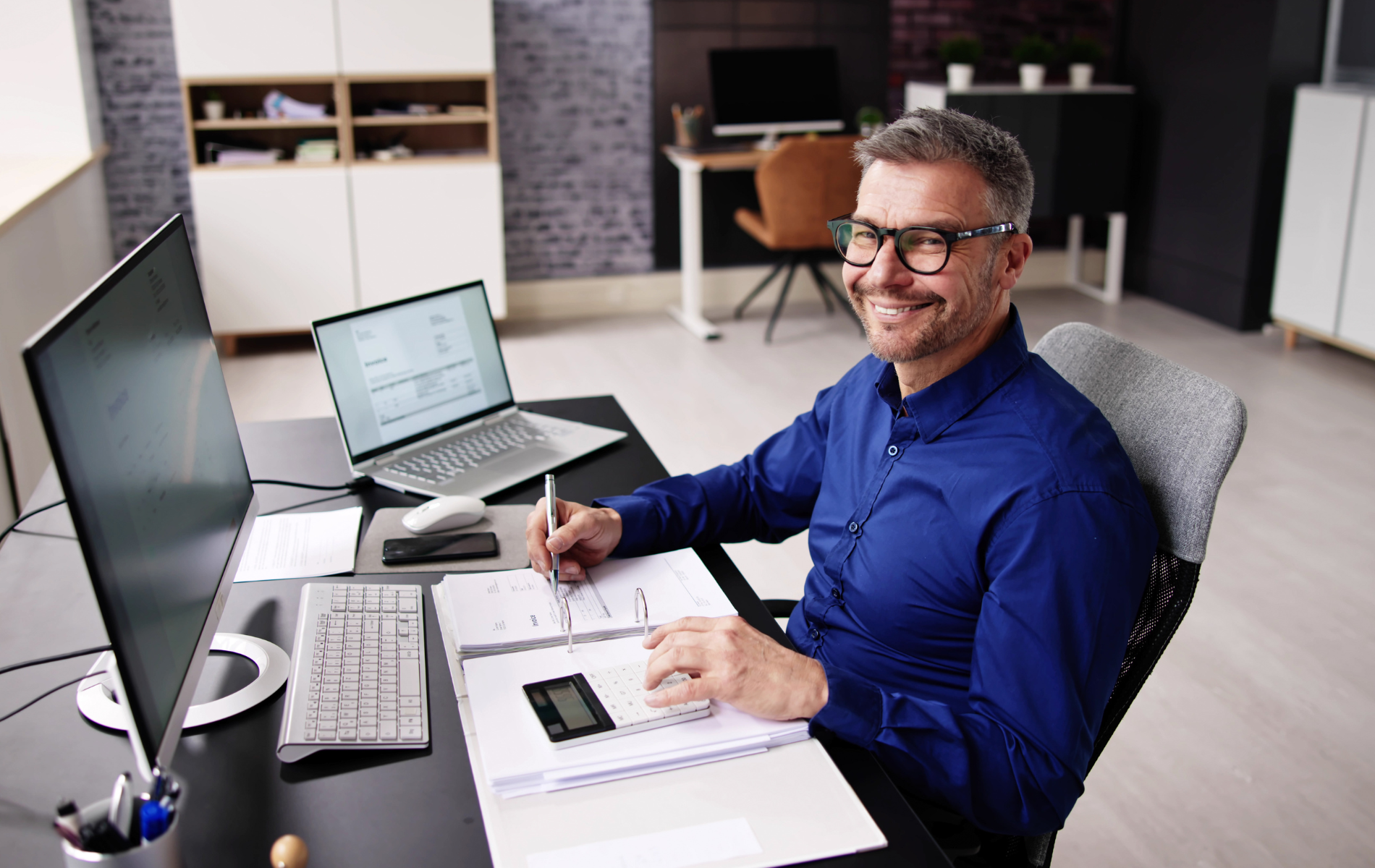 Man smiling at a desk with a laptop, notebook, and monitor in a modern office