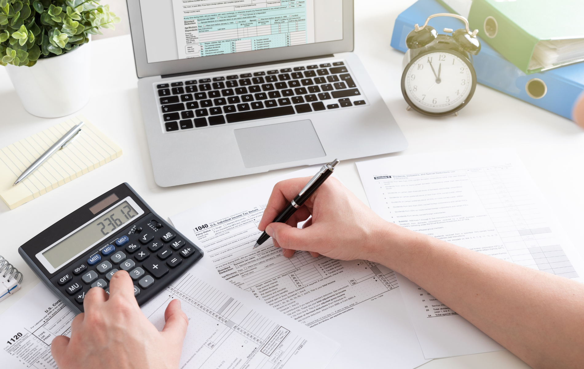 Hands calculating with a calculator while writing on paperwork beside a laptop and alarm clock on a desk