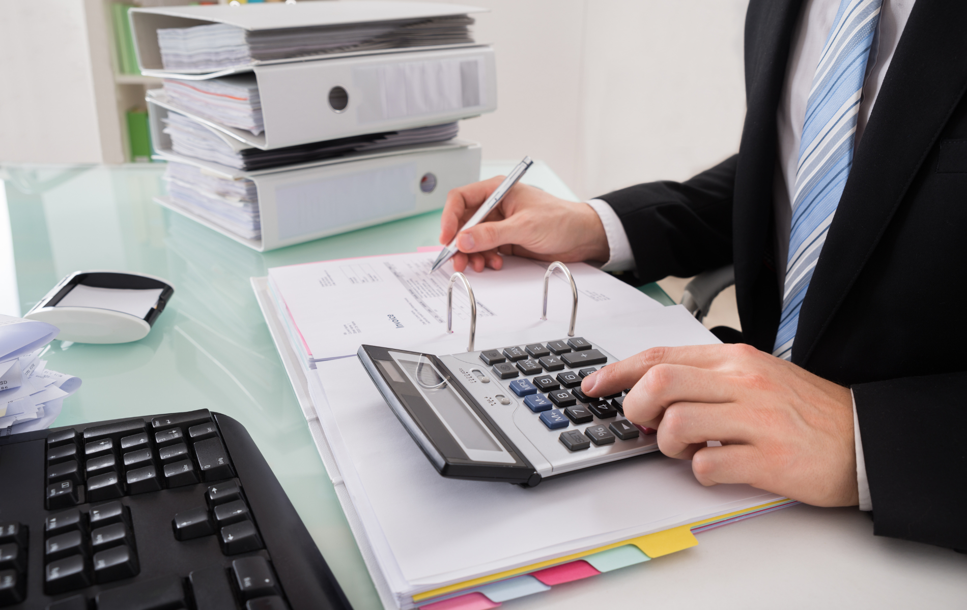 Person writing on paperwork and using a calculator at an office desk with binders nearby