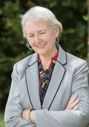 Smiling woman with short white hair in a light gray blazer, standing outdoors with arms crossed.