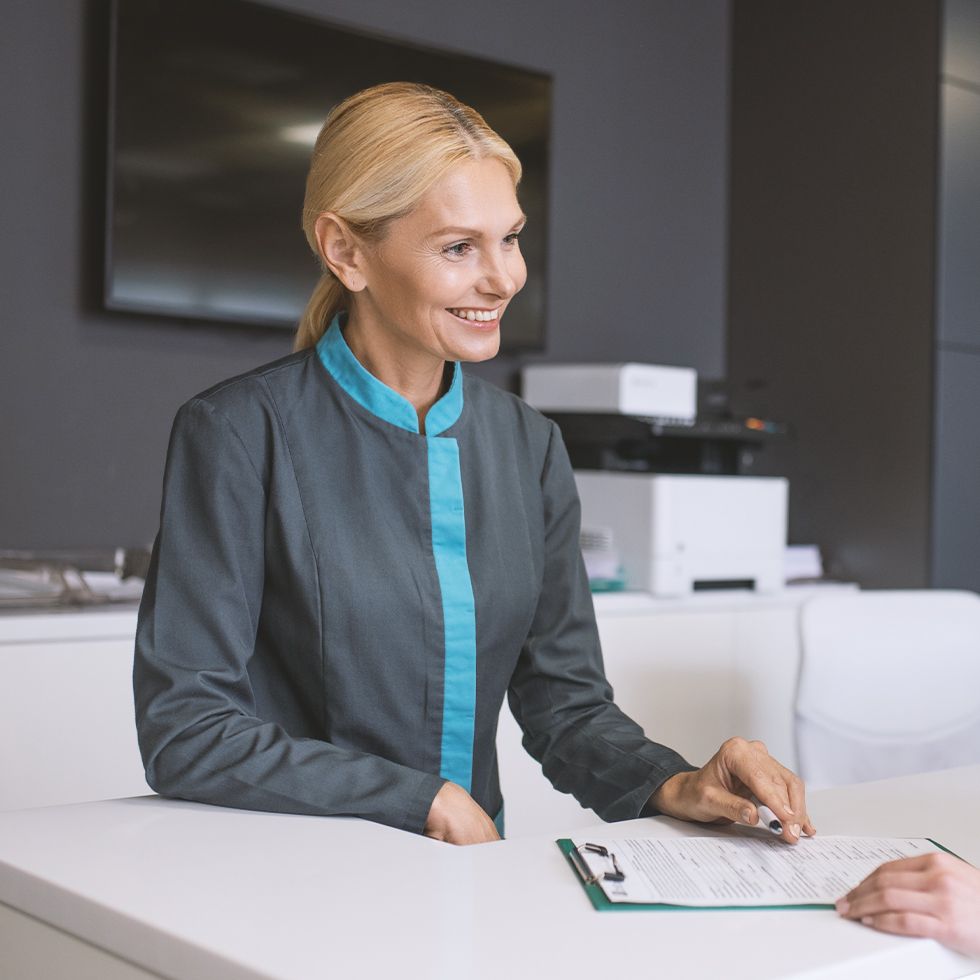 A woman is sitting at a desk with a clipboard and smiling.