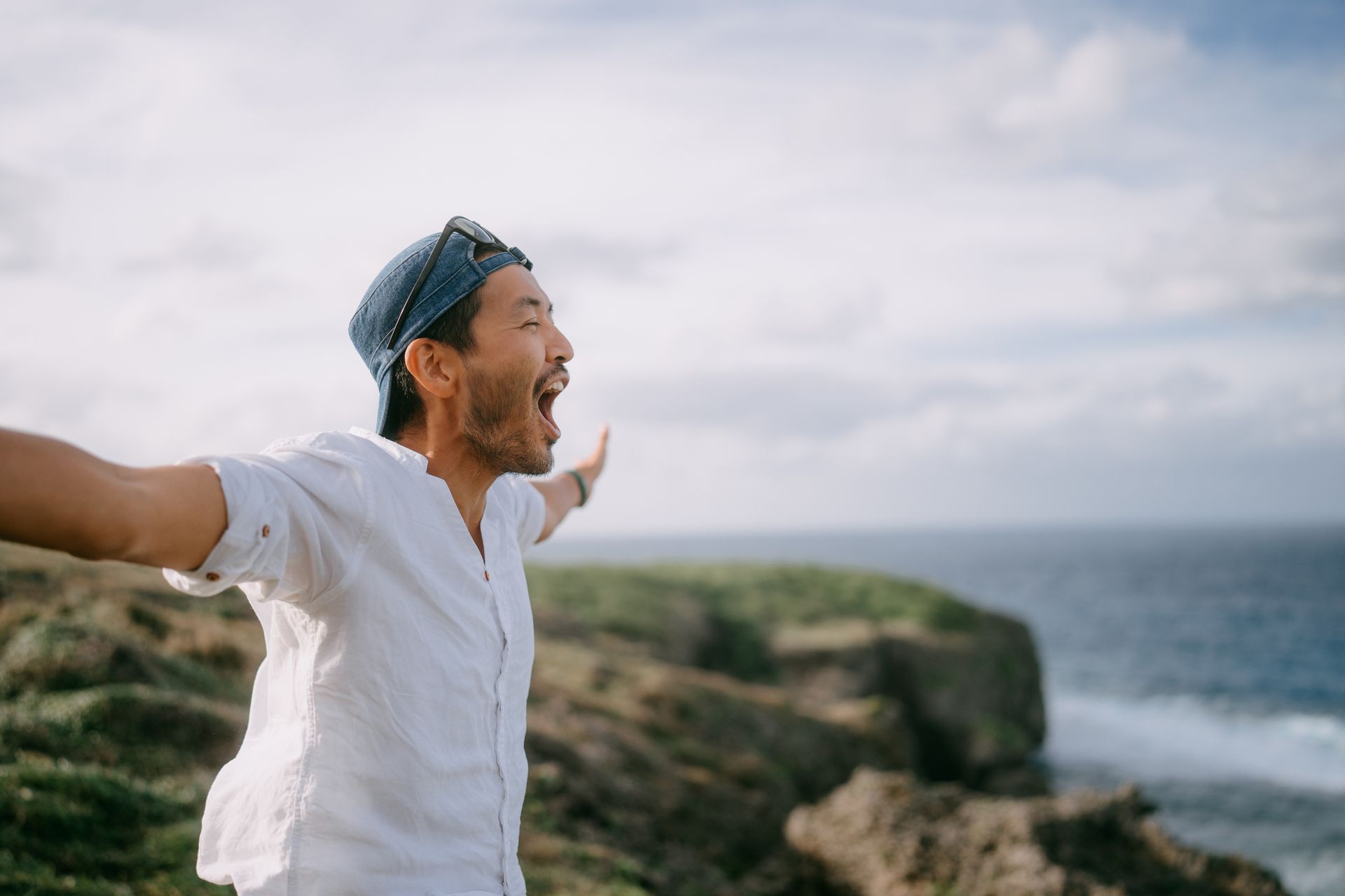 Man with arms outstretched, mouth open, standing on a cliff overlooking the ocean, joyful expression.