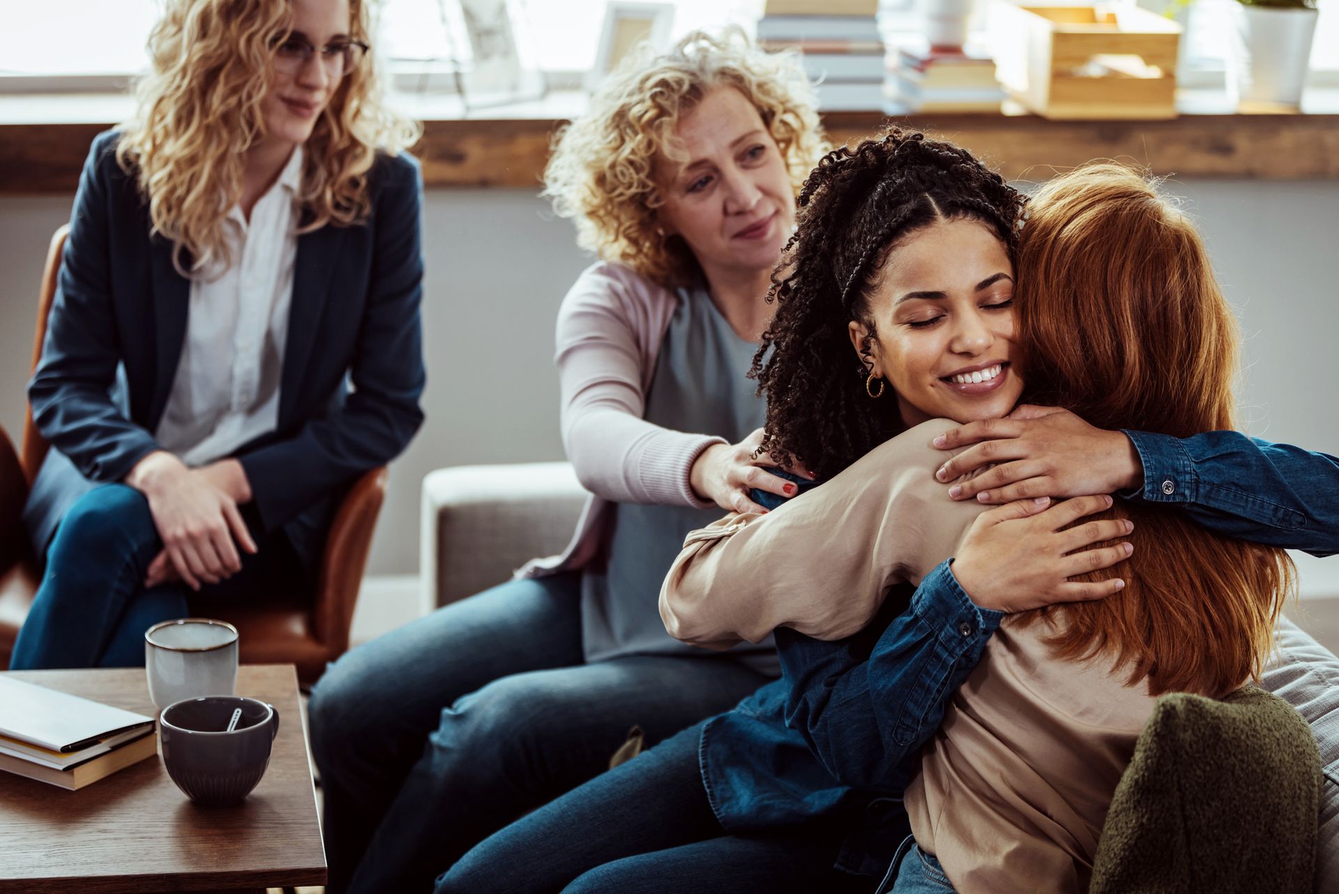 Four women in a group hug; one in a suit watches. Indoors, soft lighting, two women in the background.