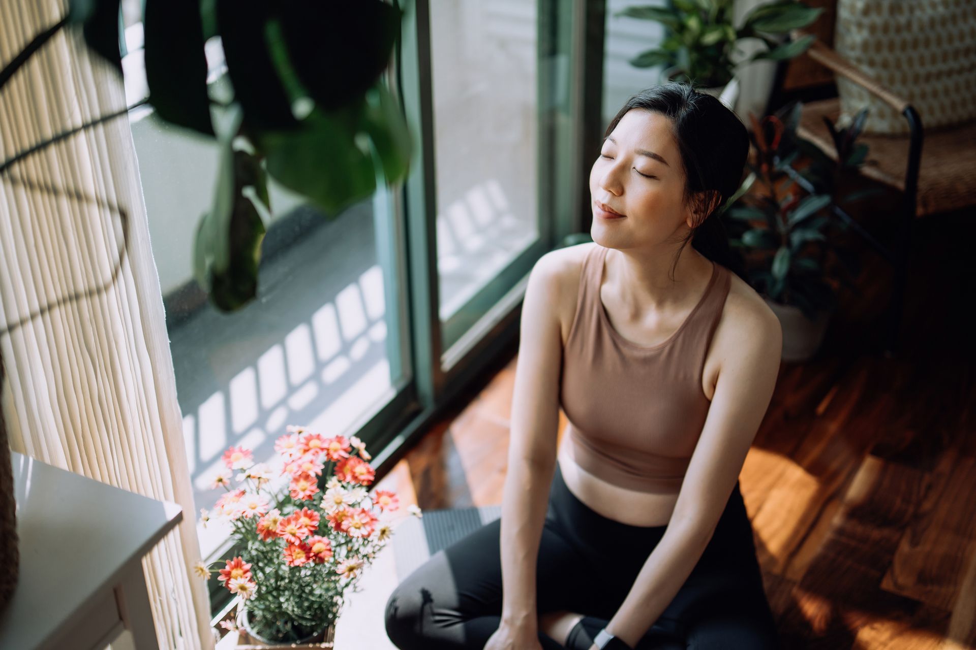 Woman in workout clothes, eyes closed, sits by a window in a sunlit room, enjoying peace.