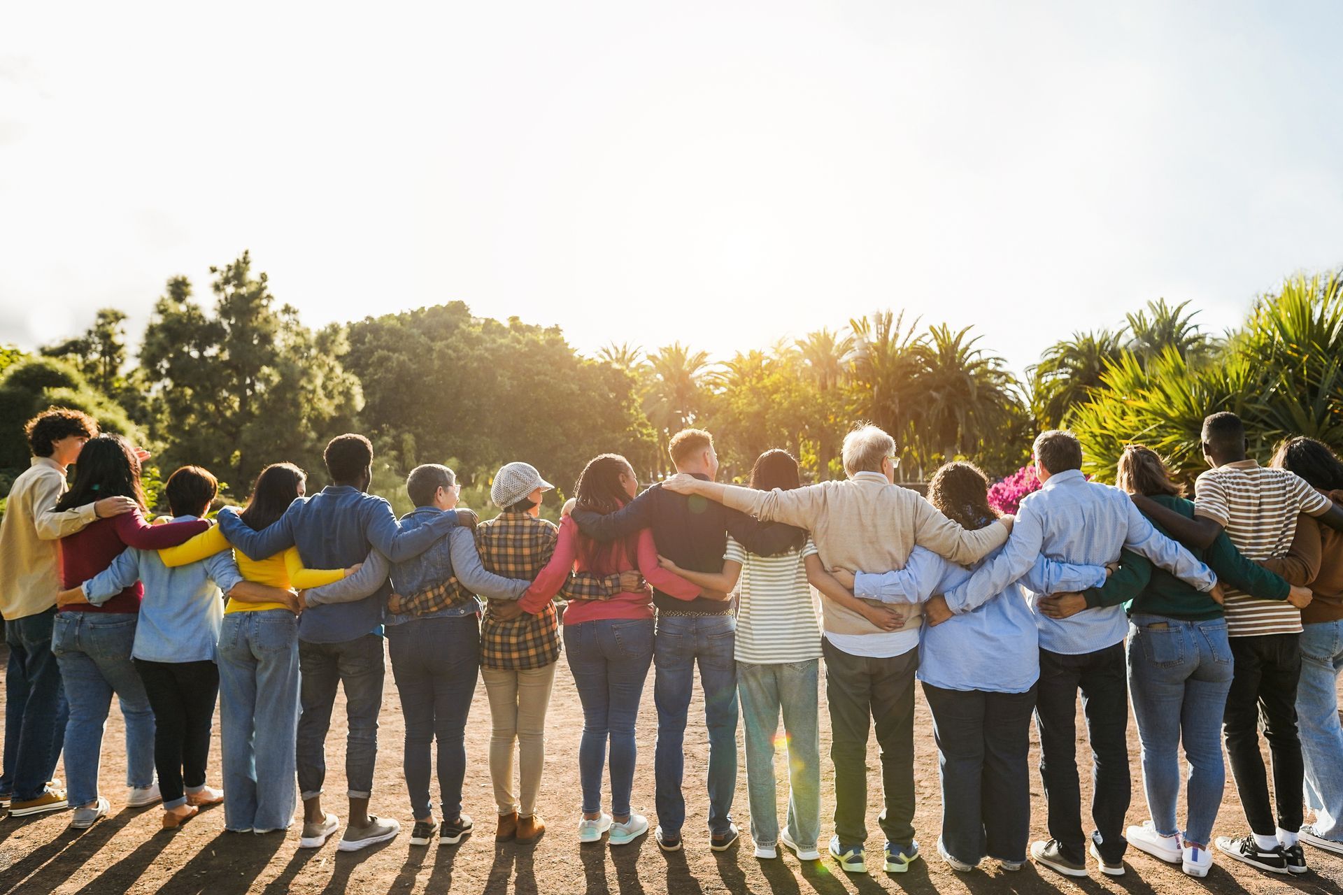 Group of diverse people with arms around each other, standing in sunlight in a park.