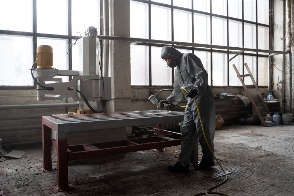 A man in a protective suit is spray painting a table in a factory.