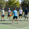 Five people on an outdoor tennis court, one in blue facing four others in a group drill.