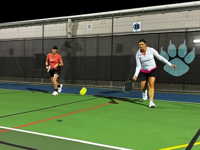 Two players rally on an indoor pickleball court, one in white and one in orange.